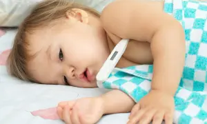Małe dziecko z termometrem pod pachą, owinięte w kocyk. Wygląda na chore, być może to trzydniówka.