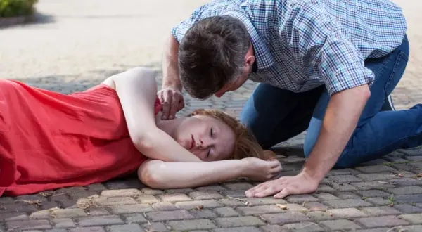 Mężczyzna sprawdza puls u nieprzytomnej kobiety leżącej na boku na chodniku.